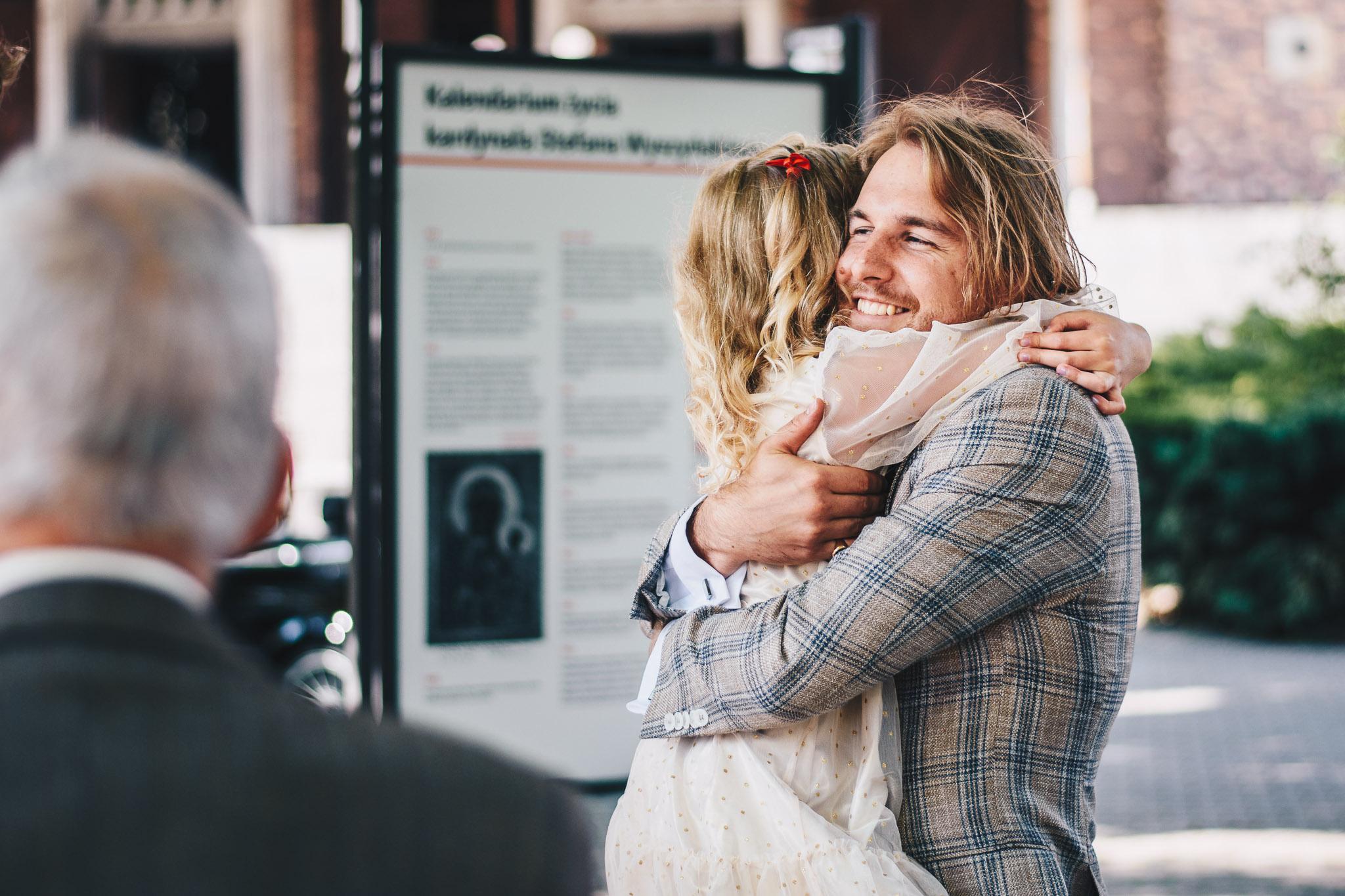 Pan młody w kraciastym garniturze trzyma na rękach małą dziewczynkę i mocno ją przytula.