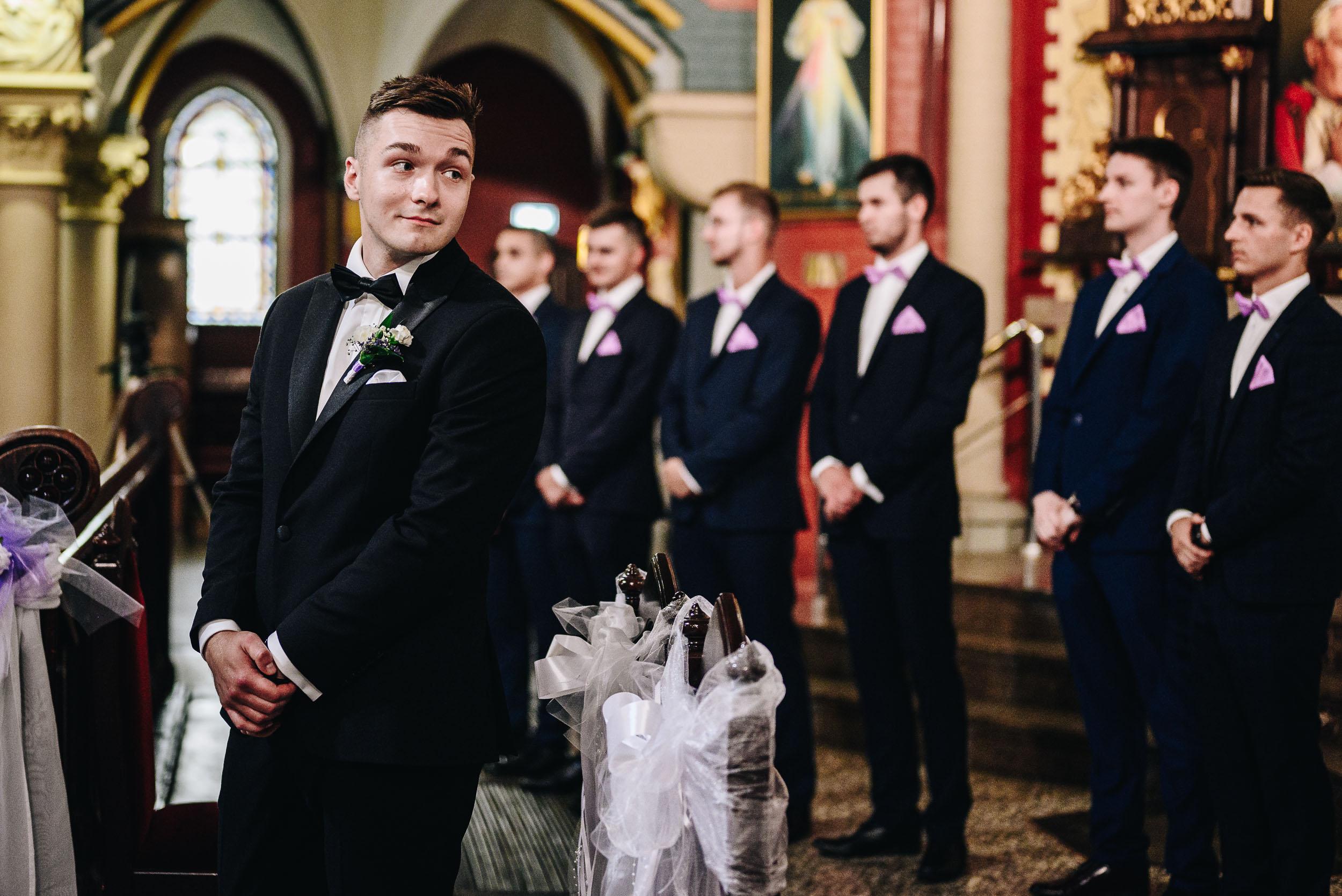 Pan młody stoi przy ołtarzu, patrząc na wchodzącą pannę młodą. Za mężczyzną stoją jego drużbowie.