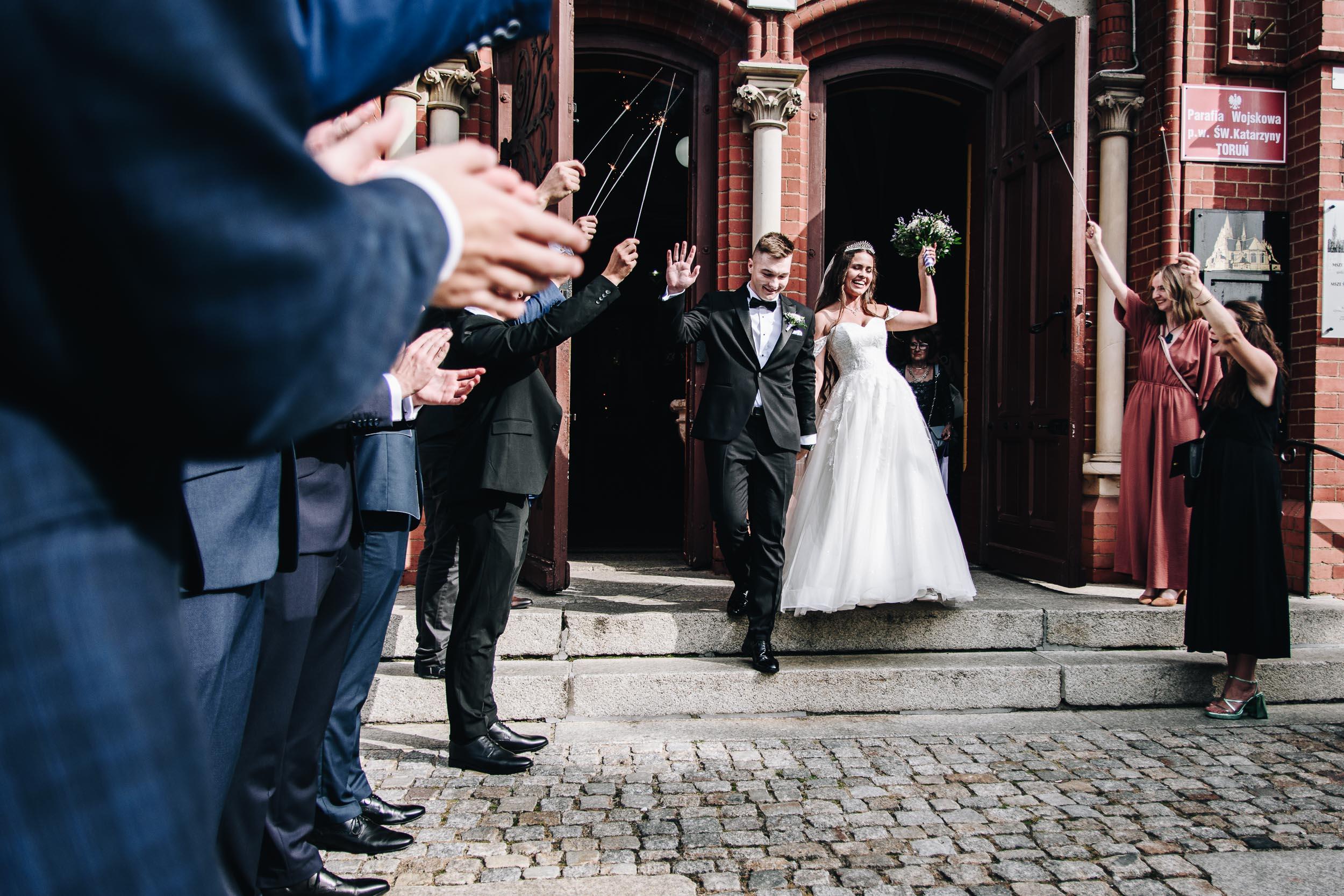 Młoda para wychodzi z kościoła w otoczeniu gości z zimnymi ogniami.