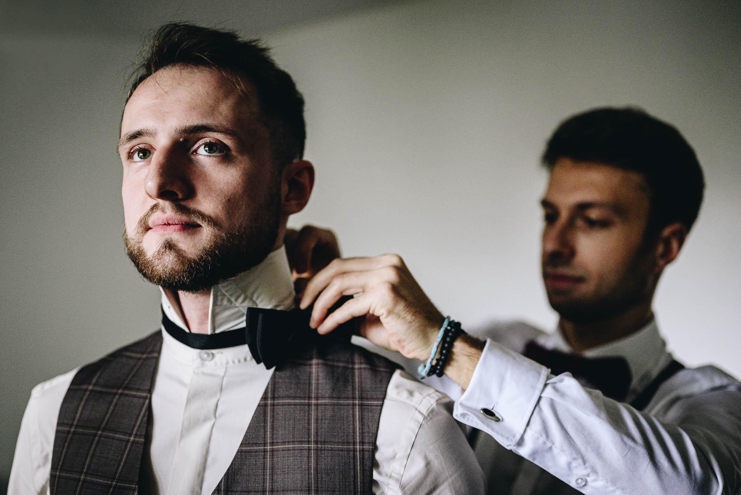 Pan młody w kamizelce i białej koszuli ma poprawianą przez świadka czarną muchę.