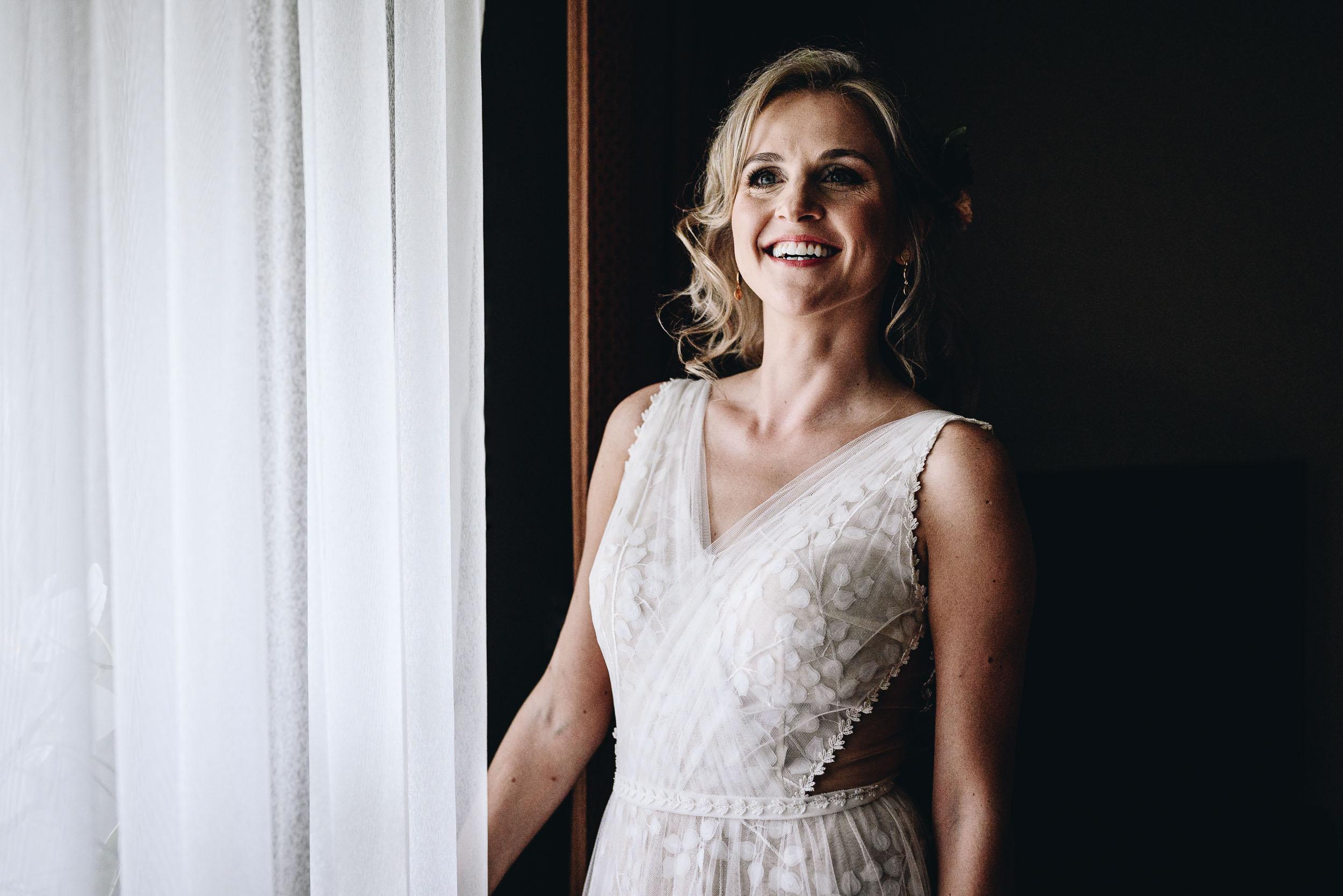 Uśmiechnięta panna młoda stoi w białej sukni przy zasłoniętym oknie.
