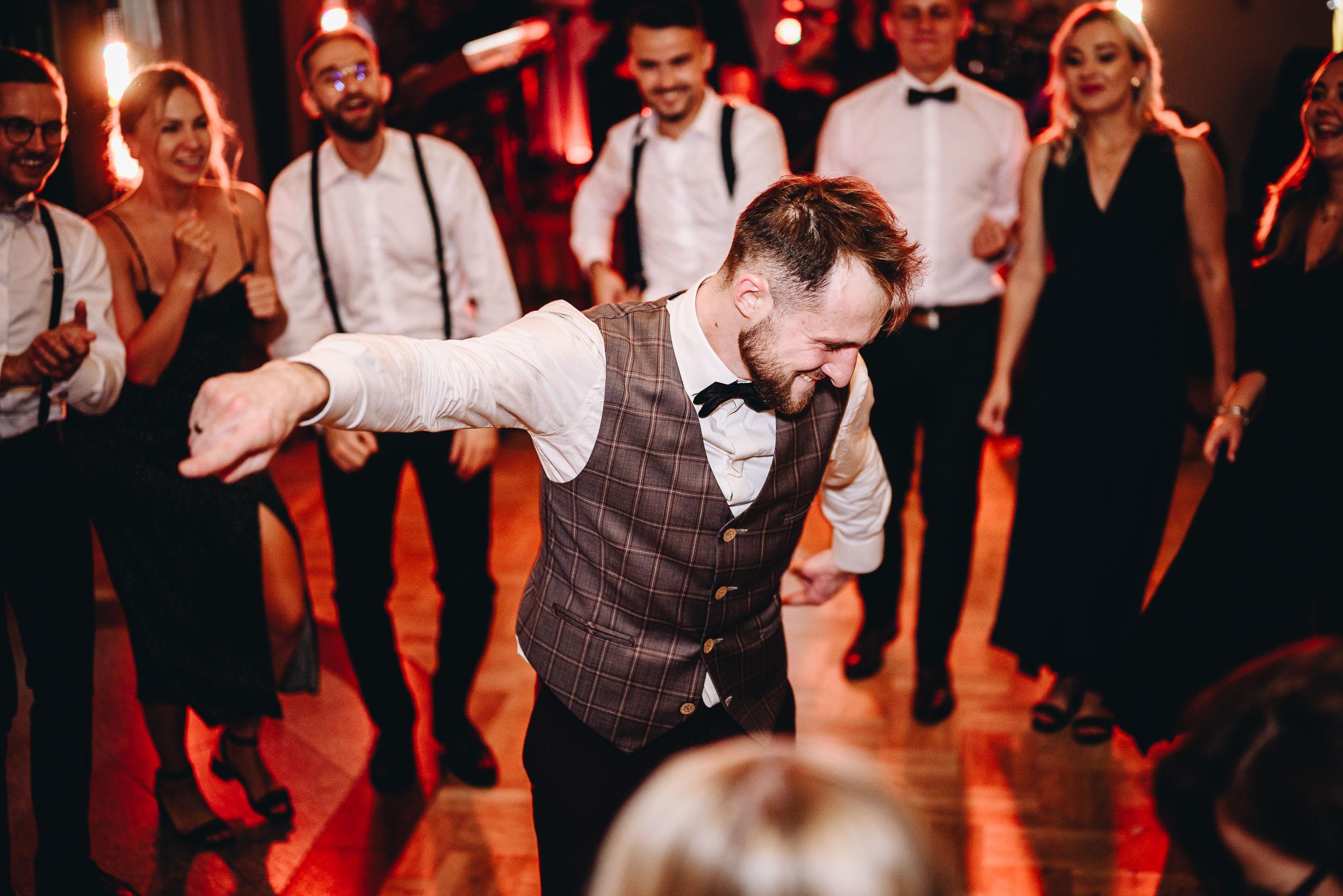 Pan młody dynamicznie tańczy solo w otoczeniu rozbawionych gości weselnych.