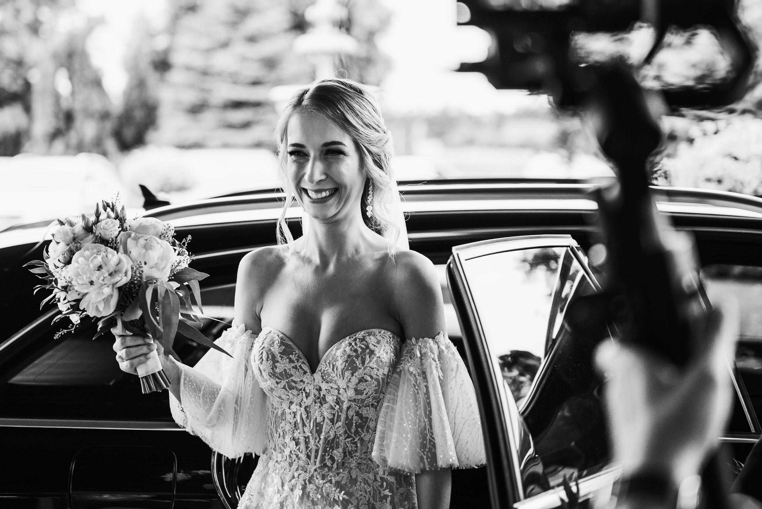 Uśmiechnięta panna młoda wychodzi z samochodu. W ręku trzyma bukiet ślubny