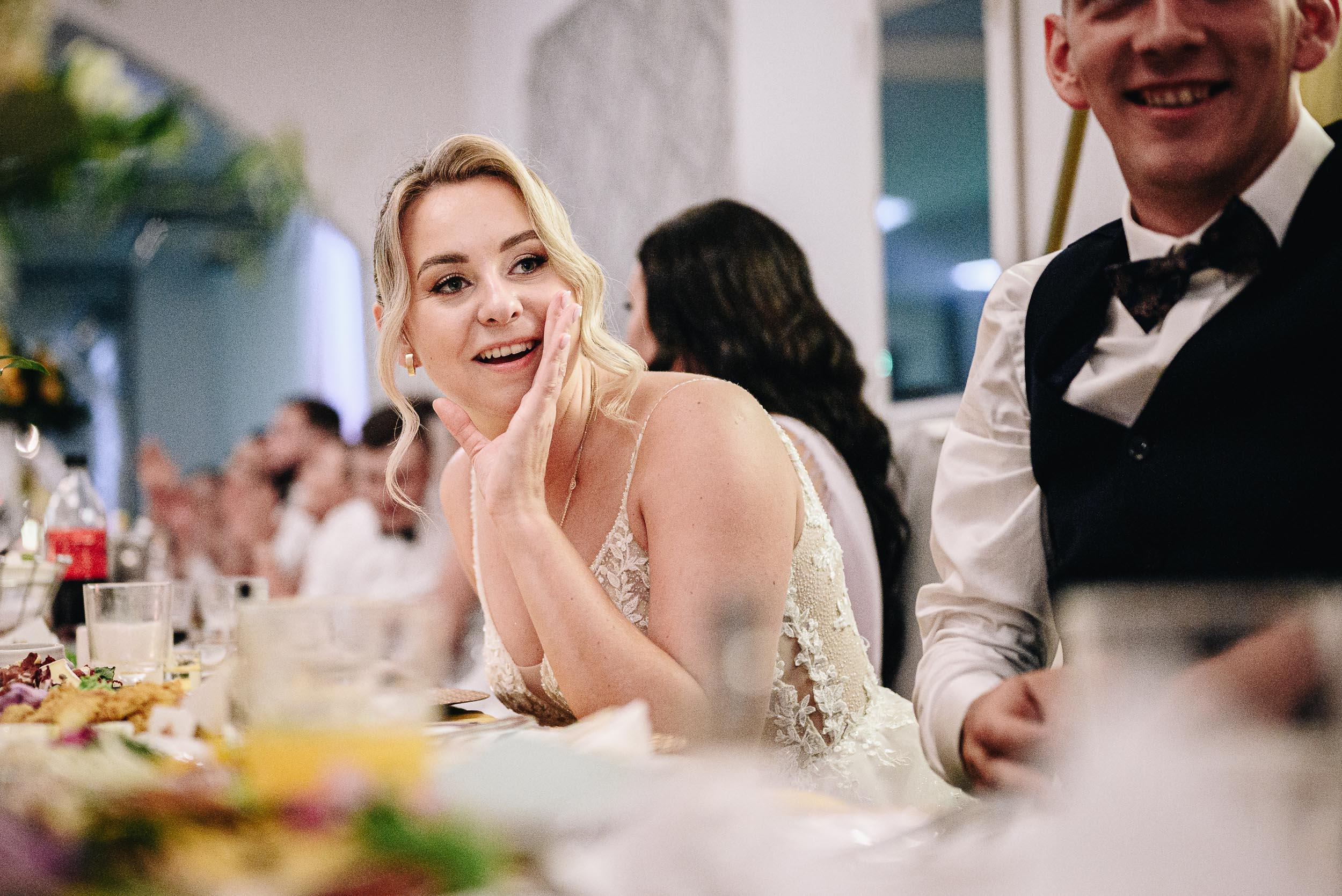 Uśmiechnięta panna młoda siedzi przy stole weselnym, i zasłaniając dłonią usta coś krzyczy.