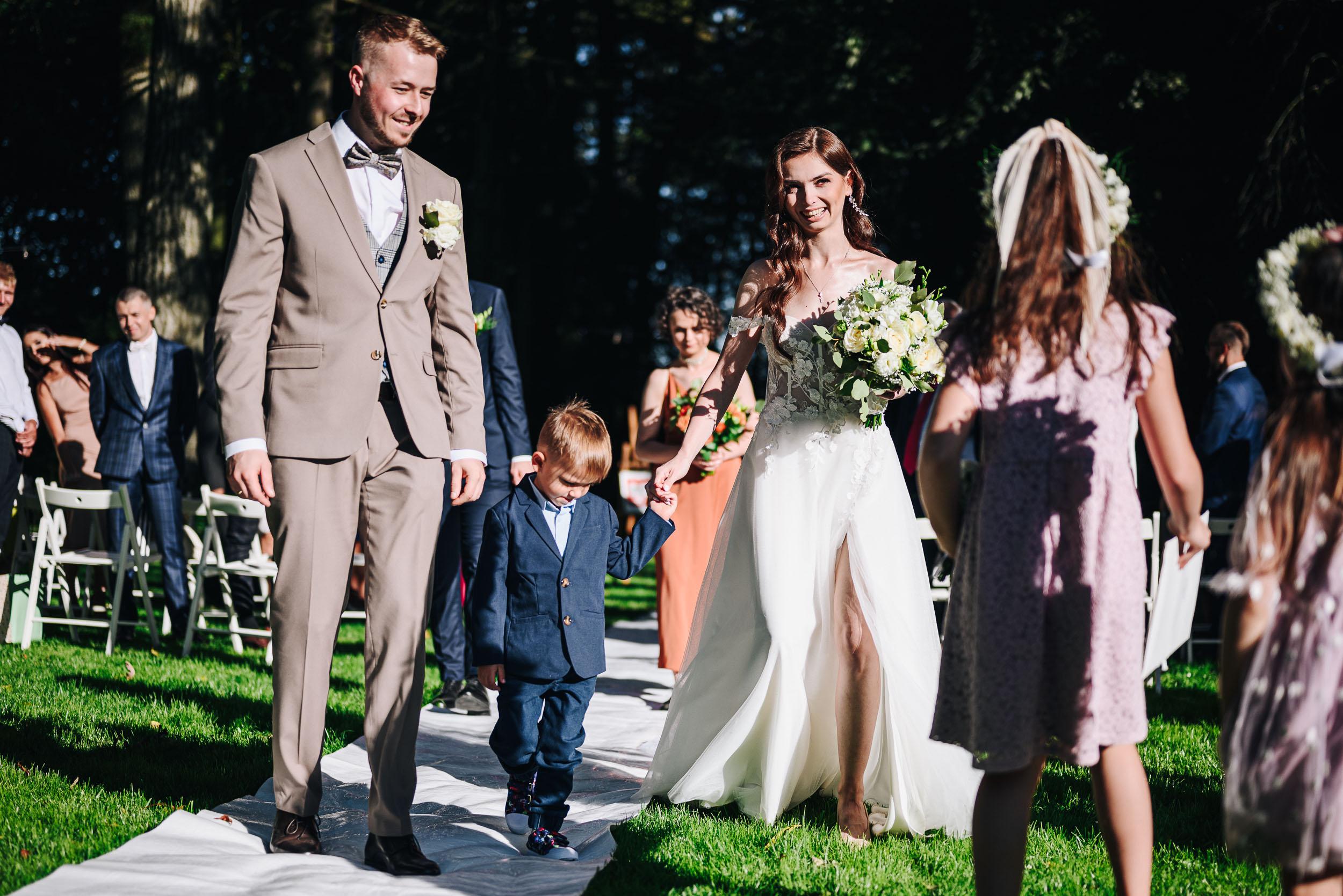 Szczęśliwa młoda para idzie w kierunku dwóch dziewczynek. Panna młoda trzyma za rękę małego chłopca.