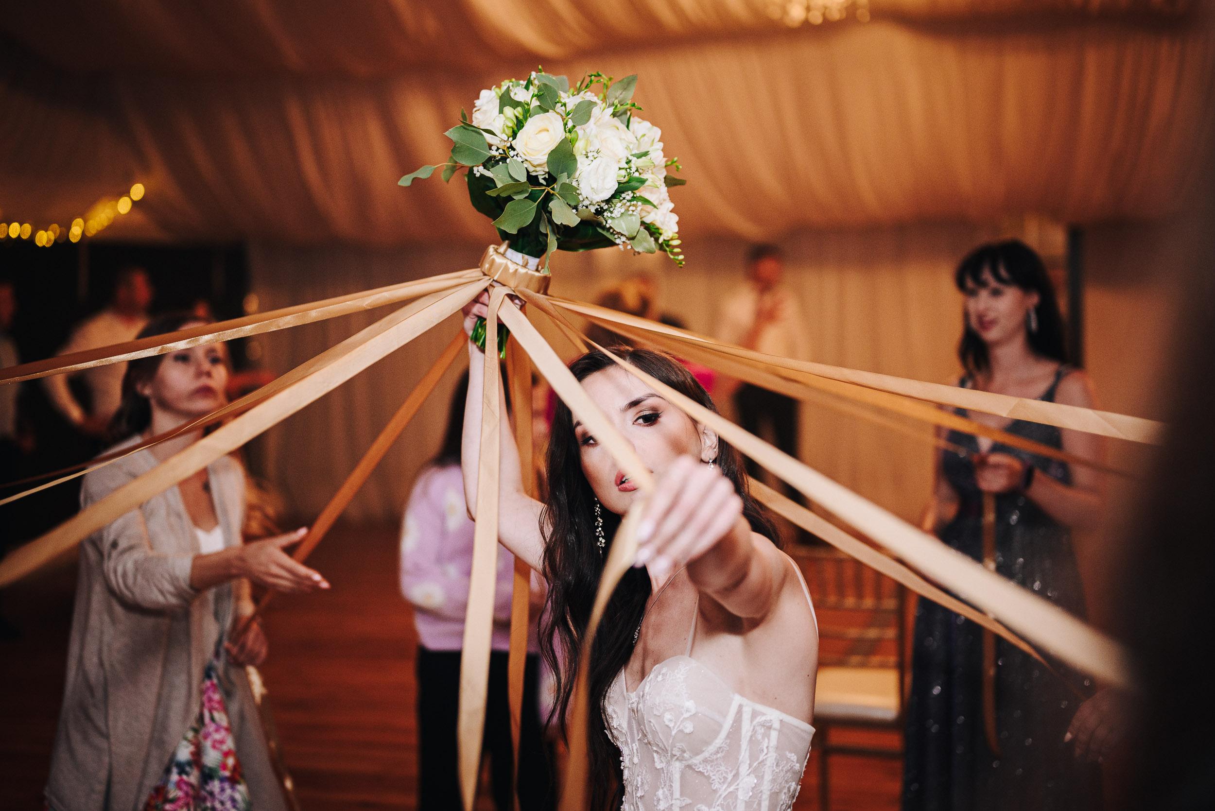 Panna młoda trzyma w ręku wstążkę od bukietu, którą próbuje podać jednej z kobiet będącej w kółku.
