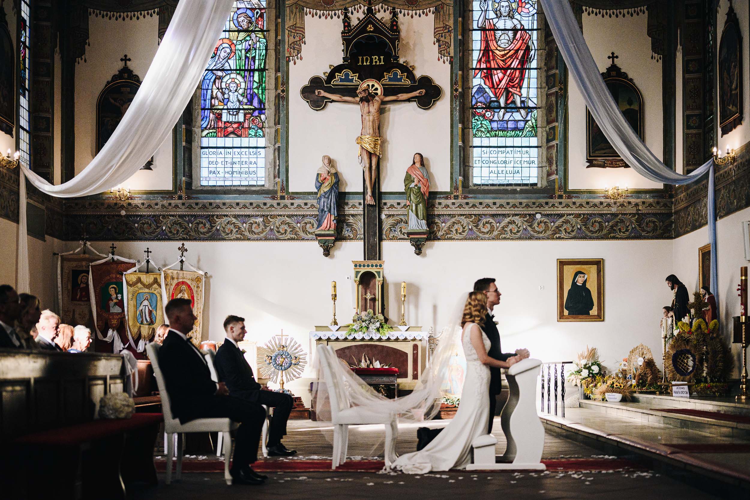 Para młoda klęczy w bogato zdobionym kościele na tle bardzo dużego krzyża