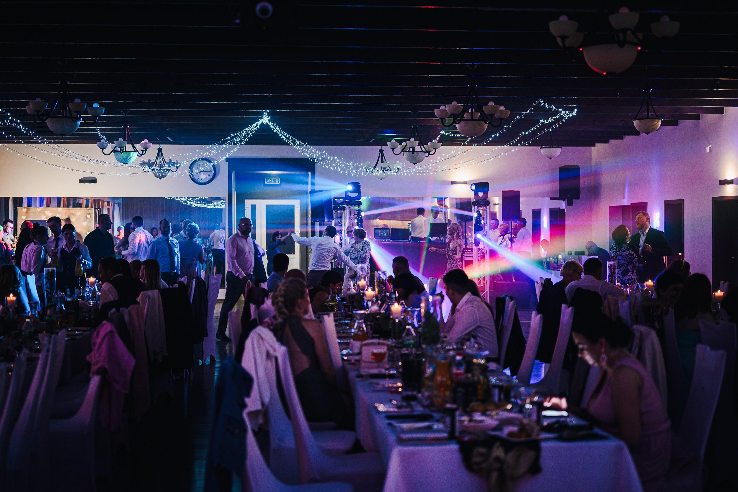 Sala weselna rozświetlona kolorowymi światła. Na parkiecie i przy stołach tłum gości.