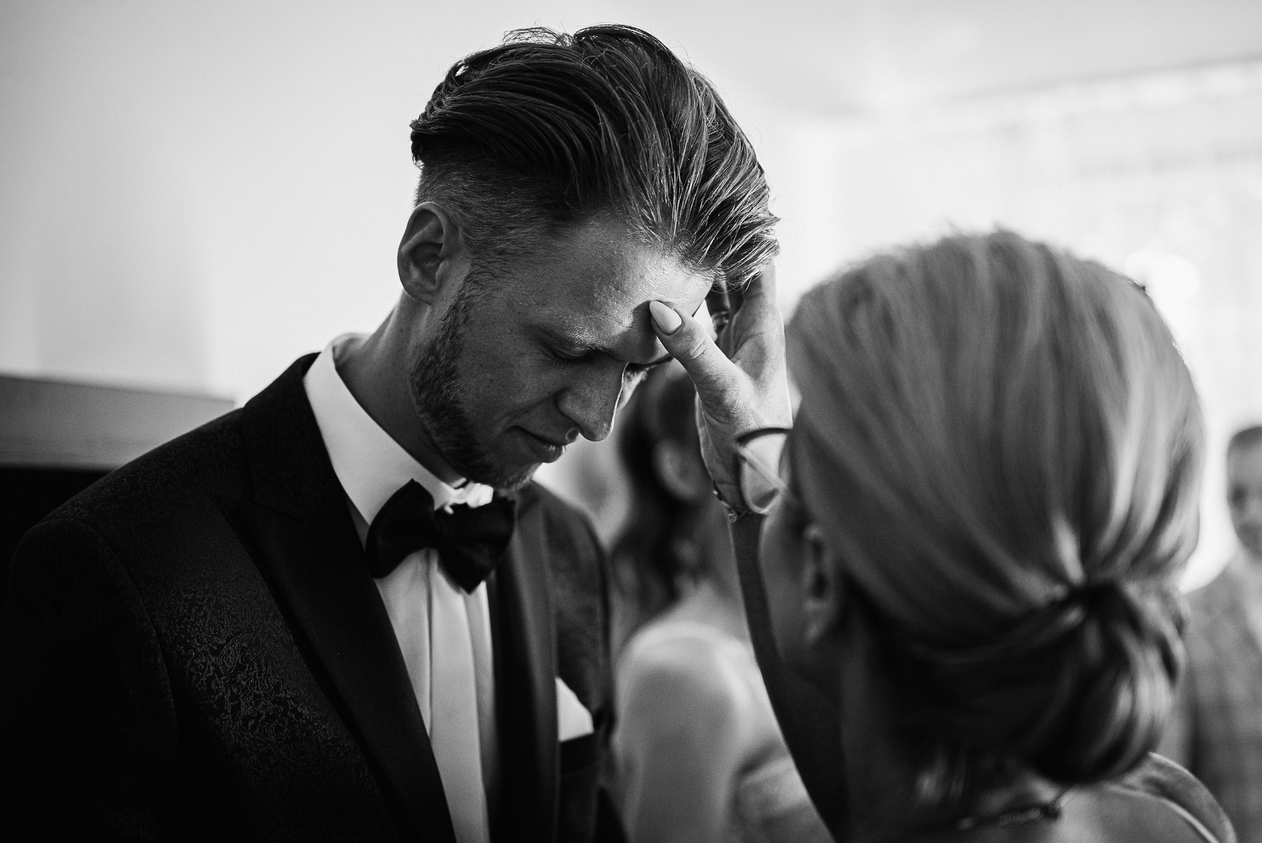 Wzruszony pan młody podczas błogosławieństwa. Kobieta wykonuje znak krzyża na jego czole.