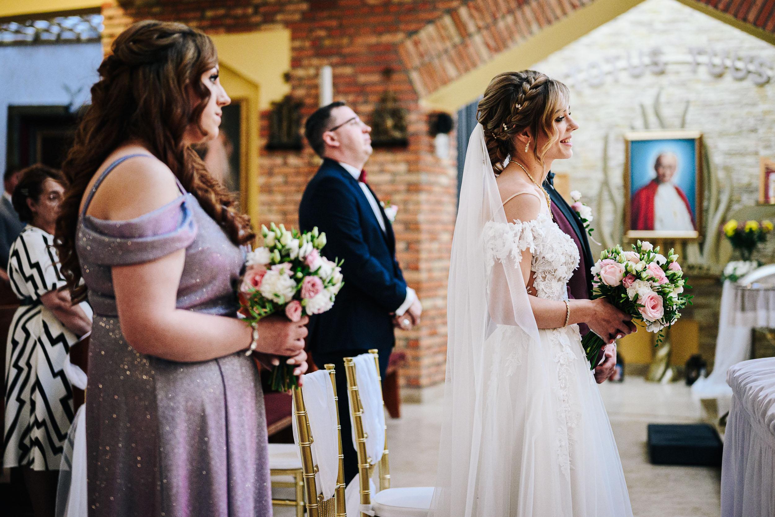 Panna młoda z bukietem i druhna w błyszczącej, fioletowej sukni stoją w kościele podczas mszy.