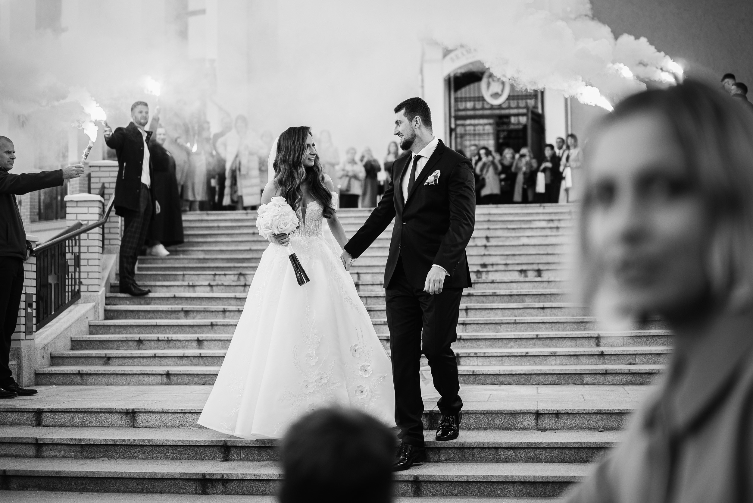 Para młoda schodzi po kamiennych schodach, otoczona gośćmi trzymającymi świece dymne.