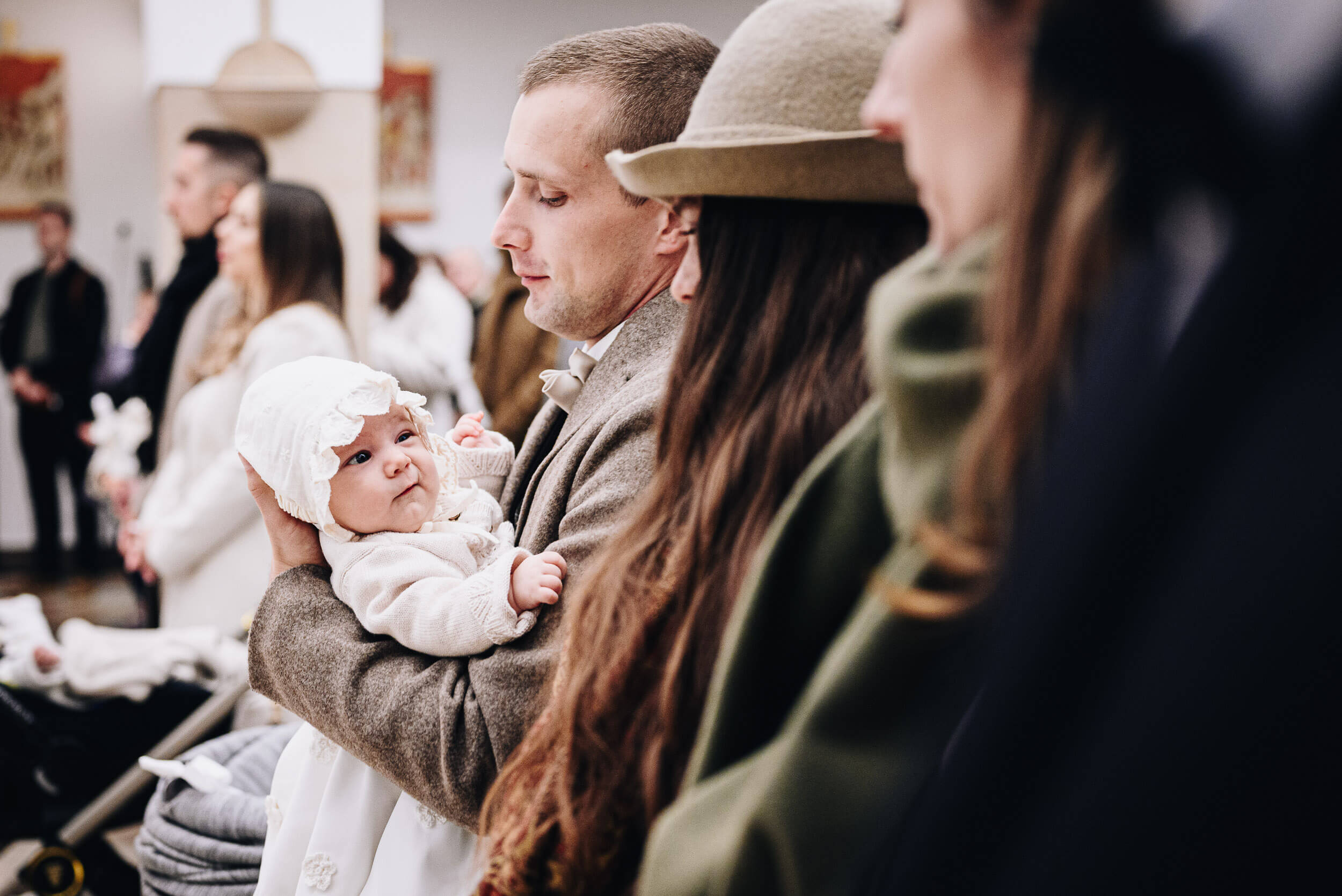 Mężczyzna w kremowym płaszczu trzyma dziewczynkę w białej sukience. Dziewczynka patrzy jednym okiem na kobietę stojącą obok.
