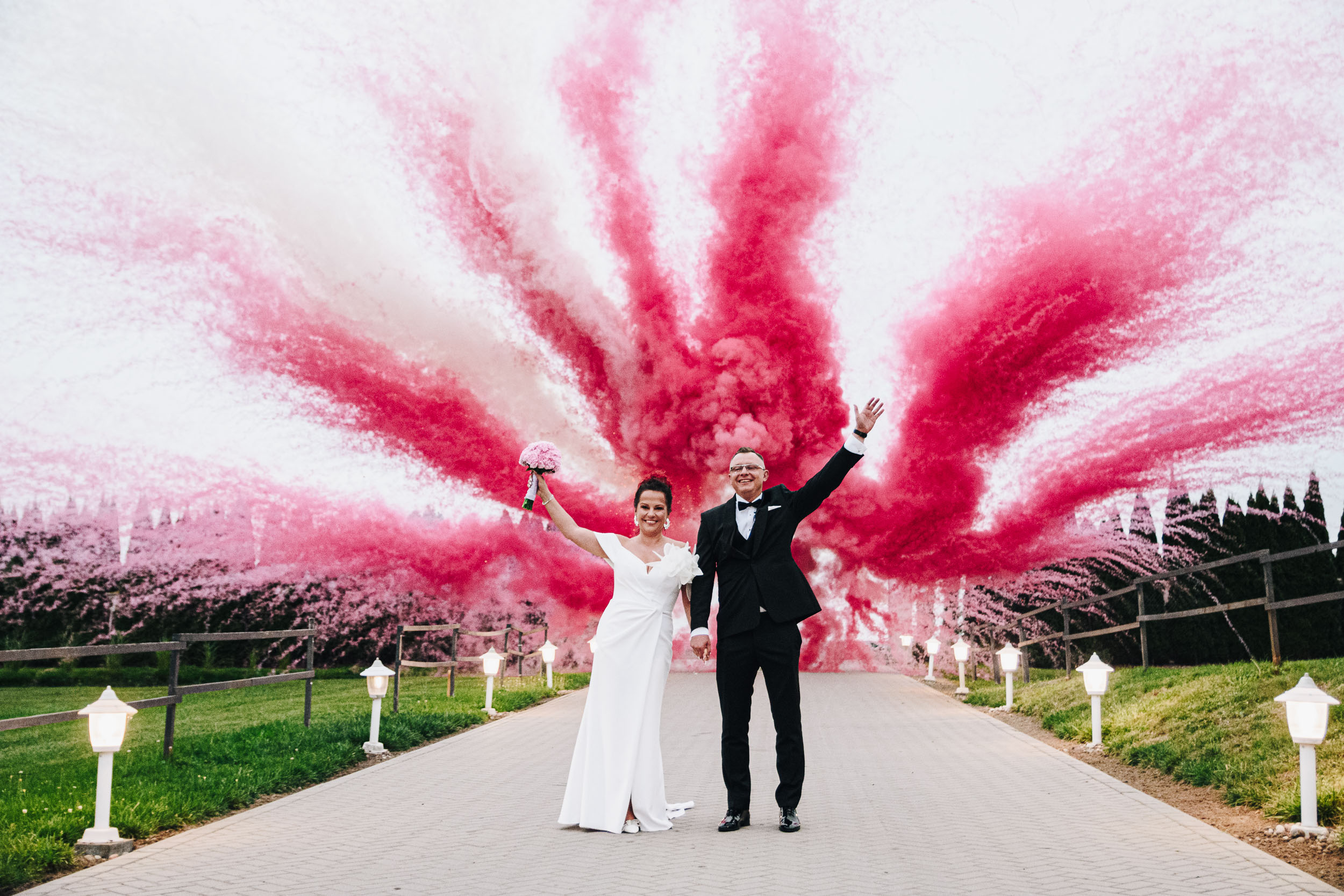 Para młoda stoi w alejce, trzyma się za ręce i macha do gości. Za zakochanymi wybuchł czerwono-biały dym.