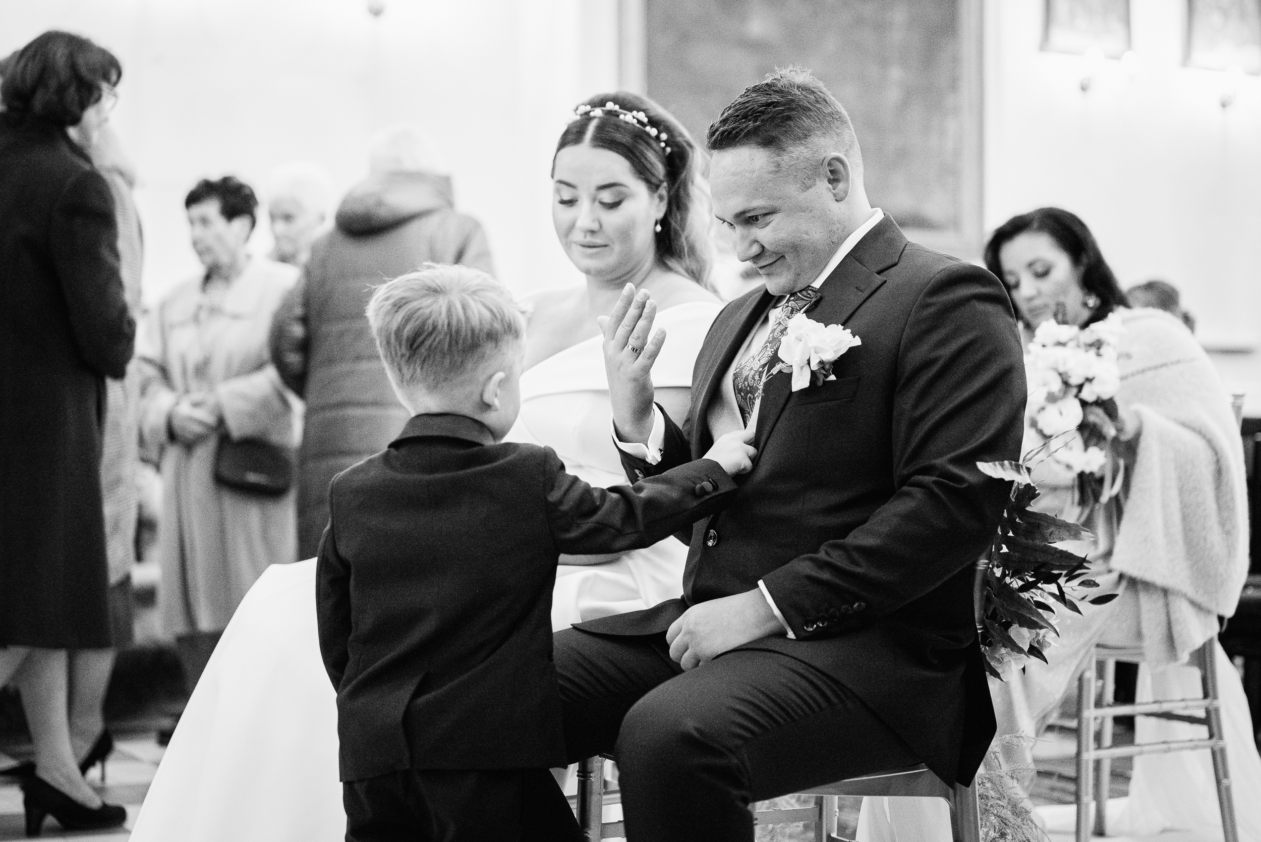 Pan młody siedzi na krześle, uśmiecha się i pokazuje chłopcu obrączkę na swoim palcu.