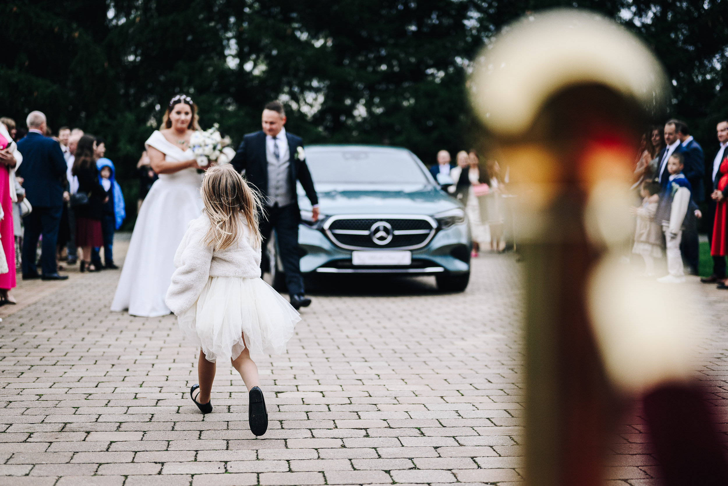 Mała dziewczynka w spódnicy biegnie w kierunku pary młodej, która wysiadła z eleganckiego samochodu