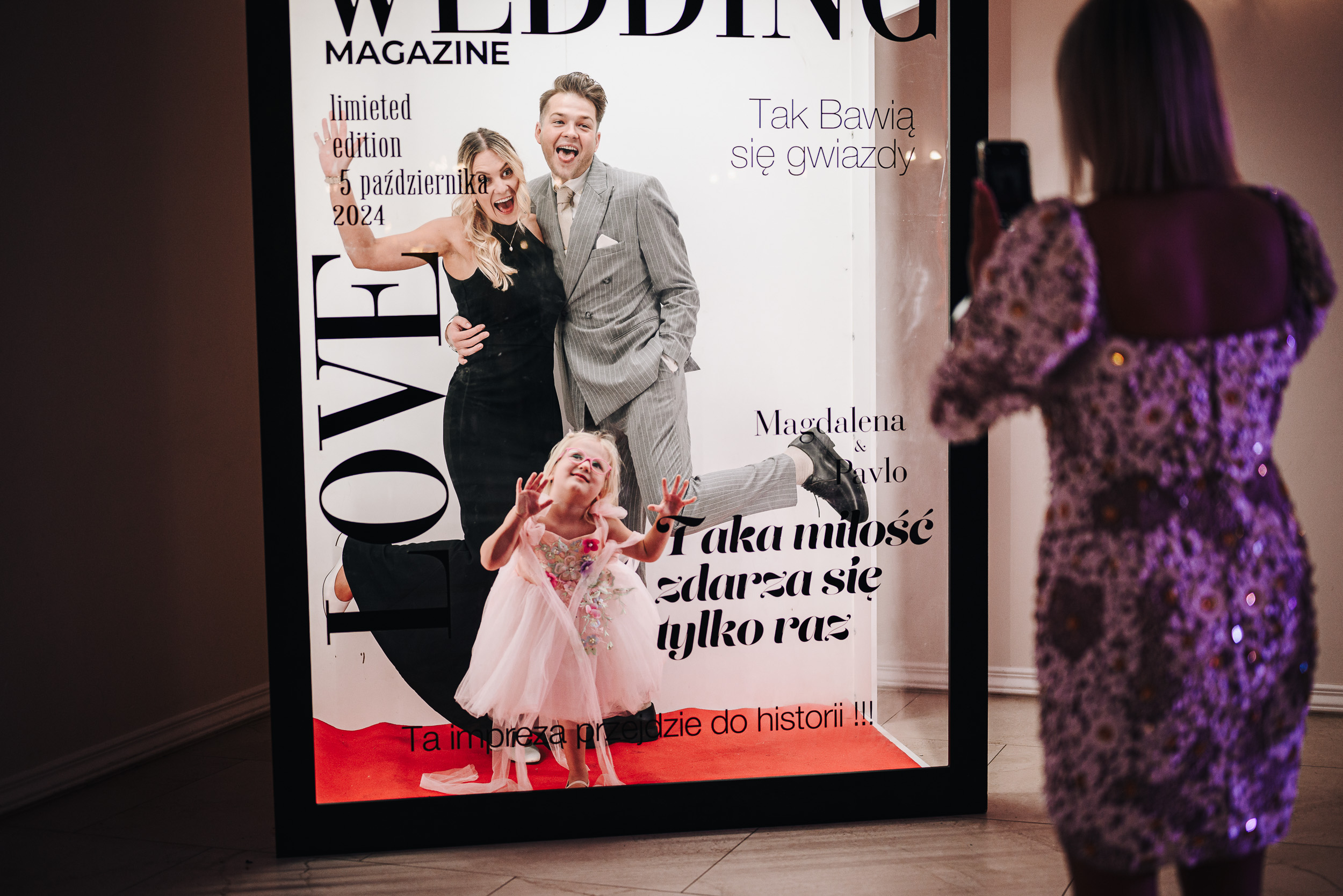 Kobieta w sukience robi zdjęcie rodzicom z dzieckiem pozującym w budce fotograficznej