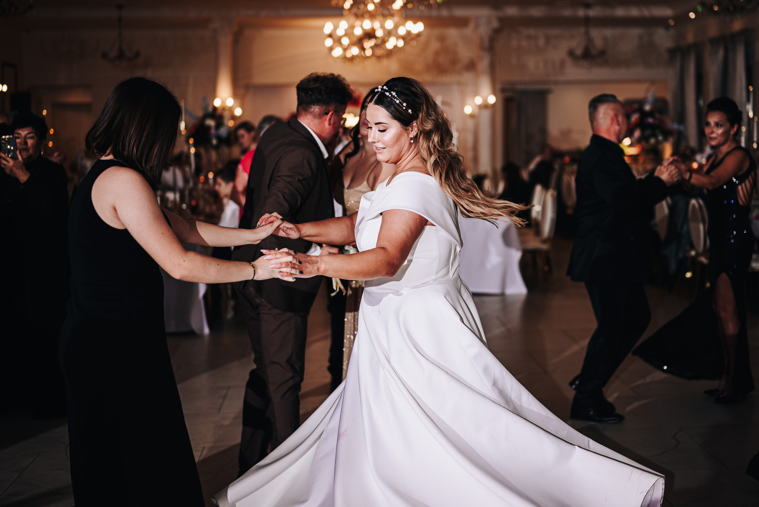 Radosna panna młoda w białej sukni wiruje w tańcu z gościem w czarnej kreacji na parkiecie sali weselnej.