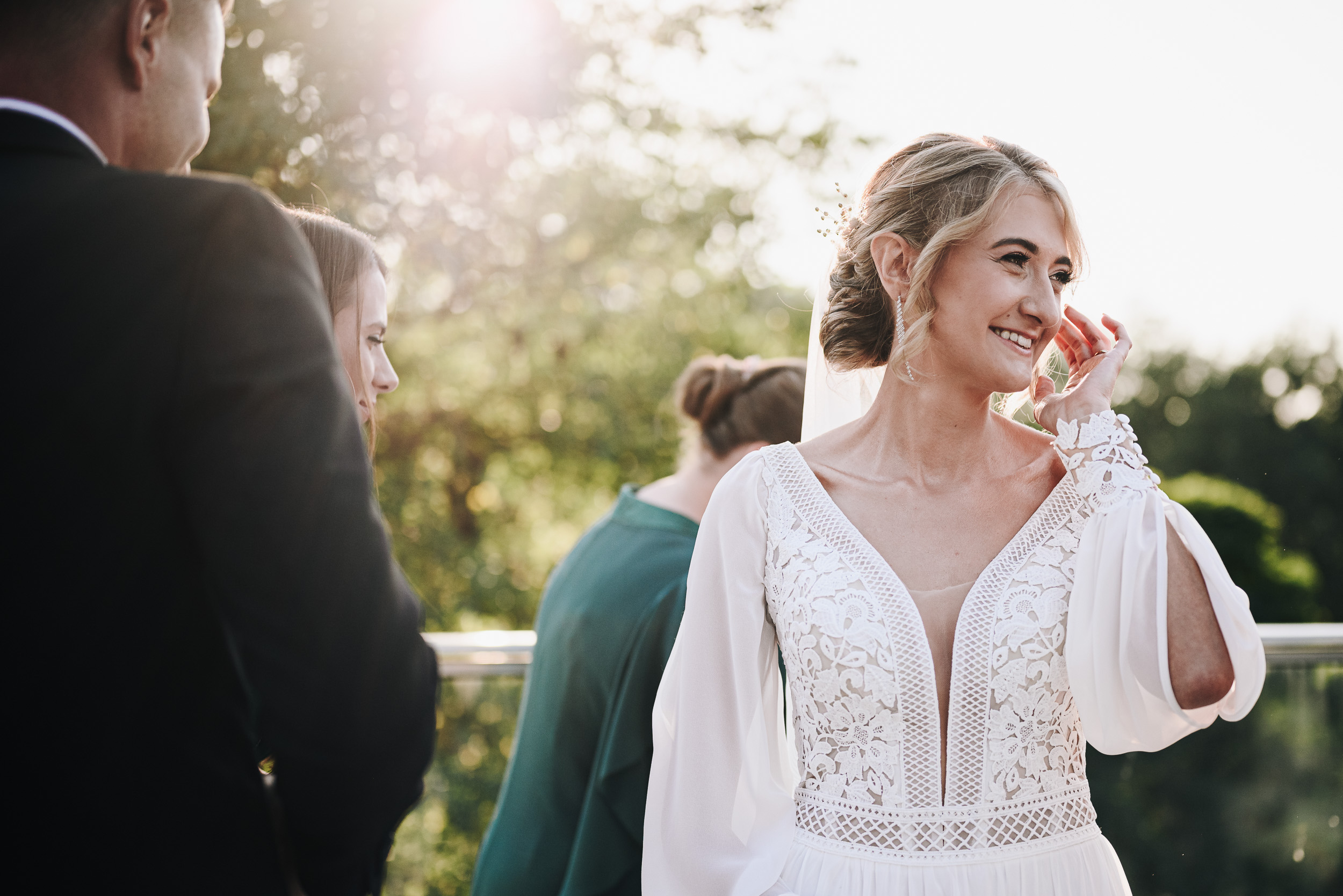 Uśmiechnięta panna młoda w koronkowej sukni poprawia włosy w promieniach słońca.