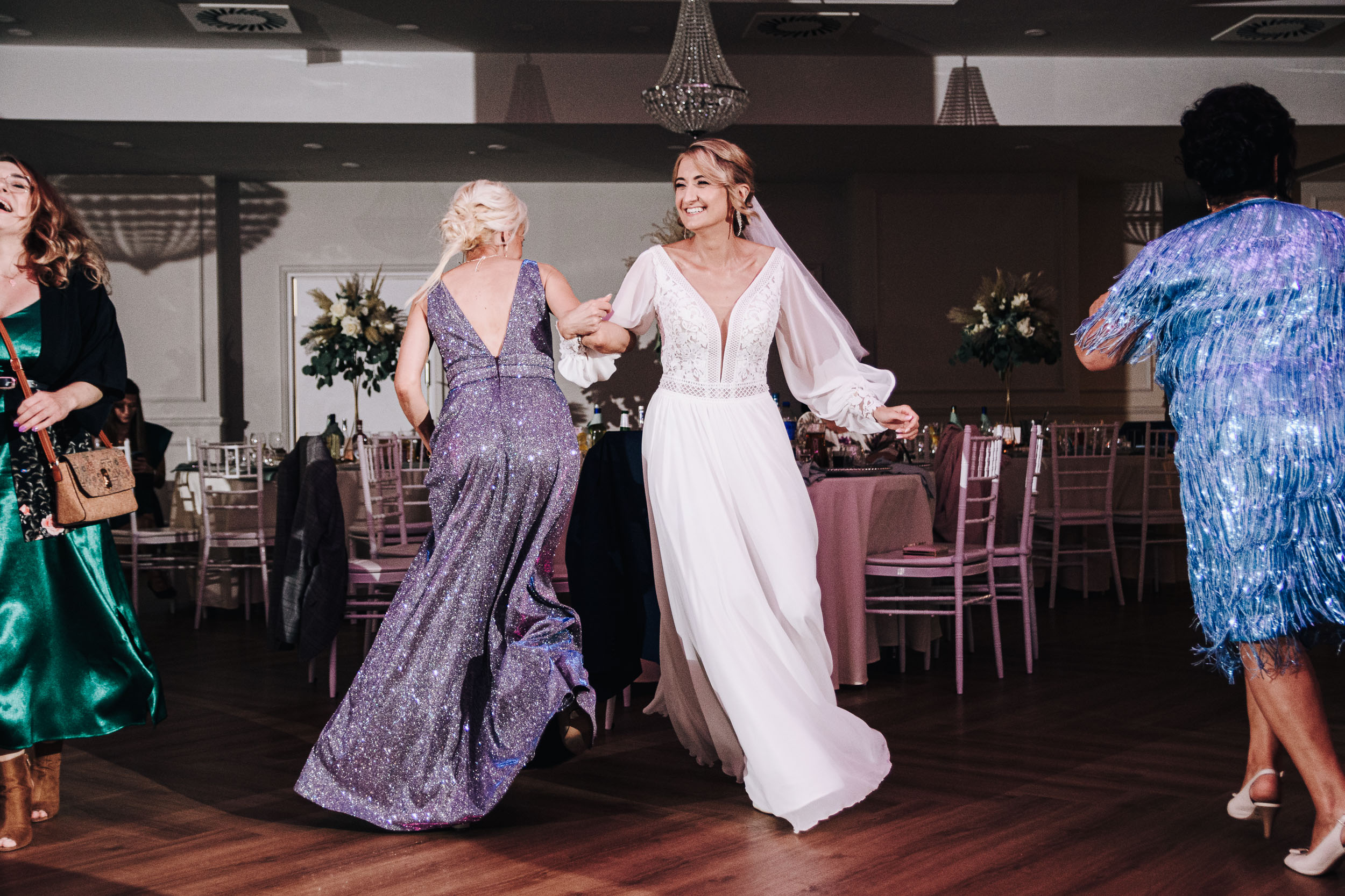Roześmiana panna młoda tańczy pod rękę z kobietą w cekinowej sukience.