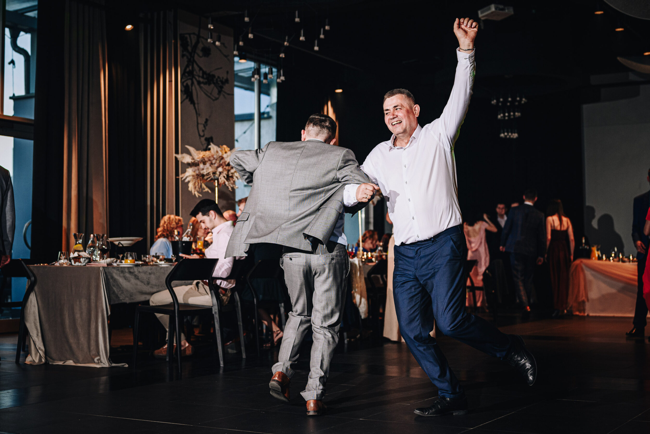 Dwóch mężczyzn tańczy pod rękę na parkiecie podczas trwania wesela. Jeden z nich trzyma rękę w górze.