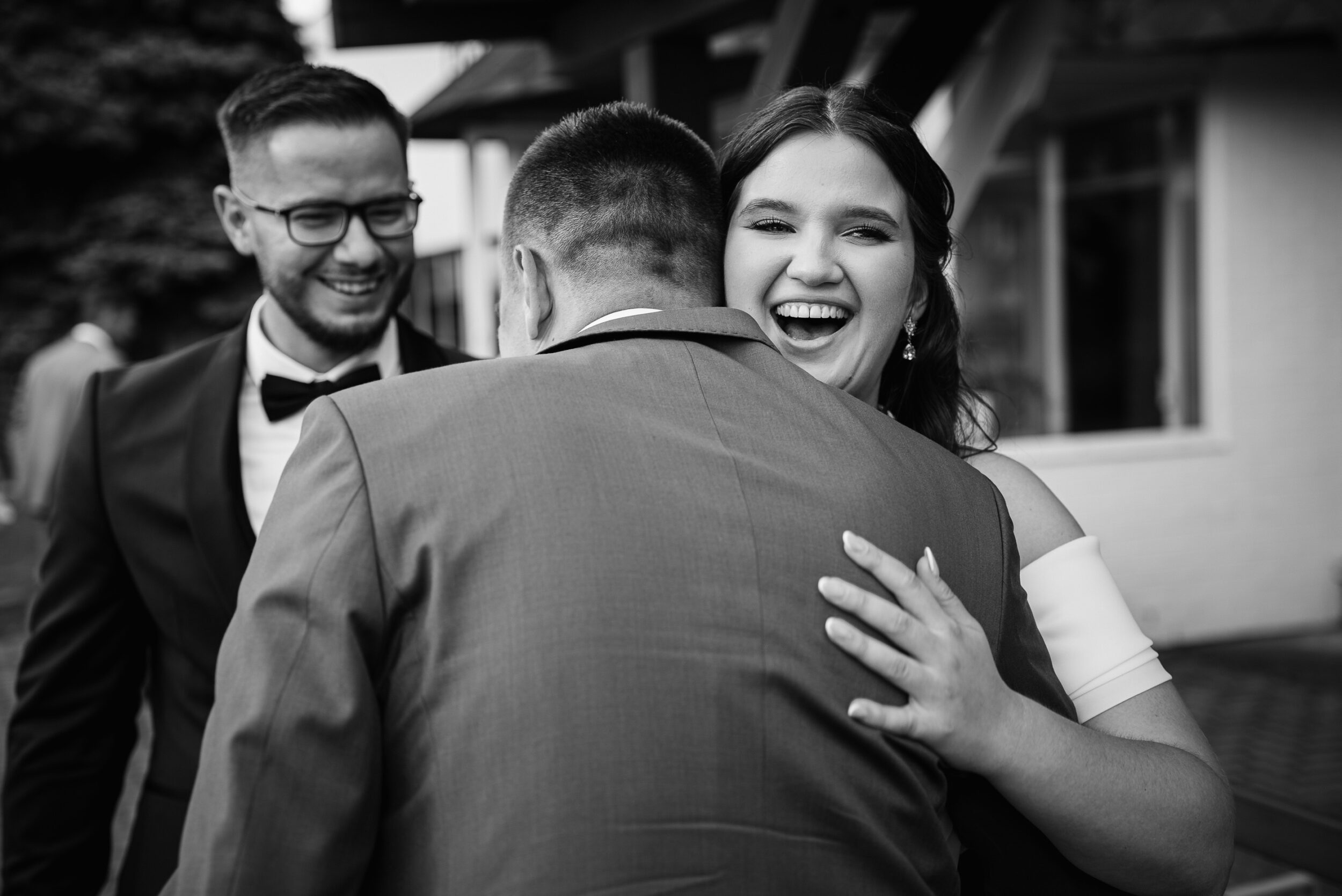 Rozpromieniona panna młoda obejmuje serdecznie mężczyznę w garniturze. W tle pan młody