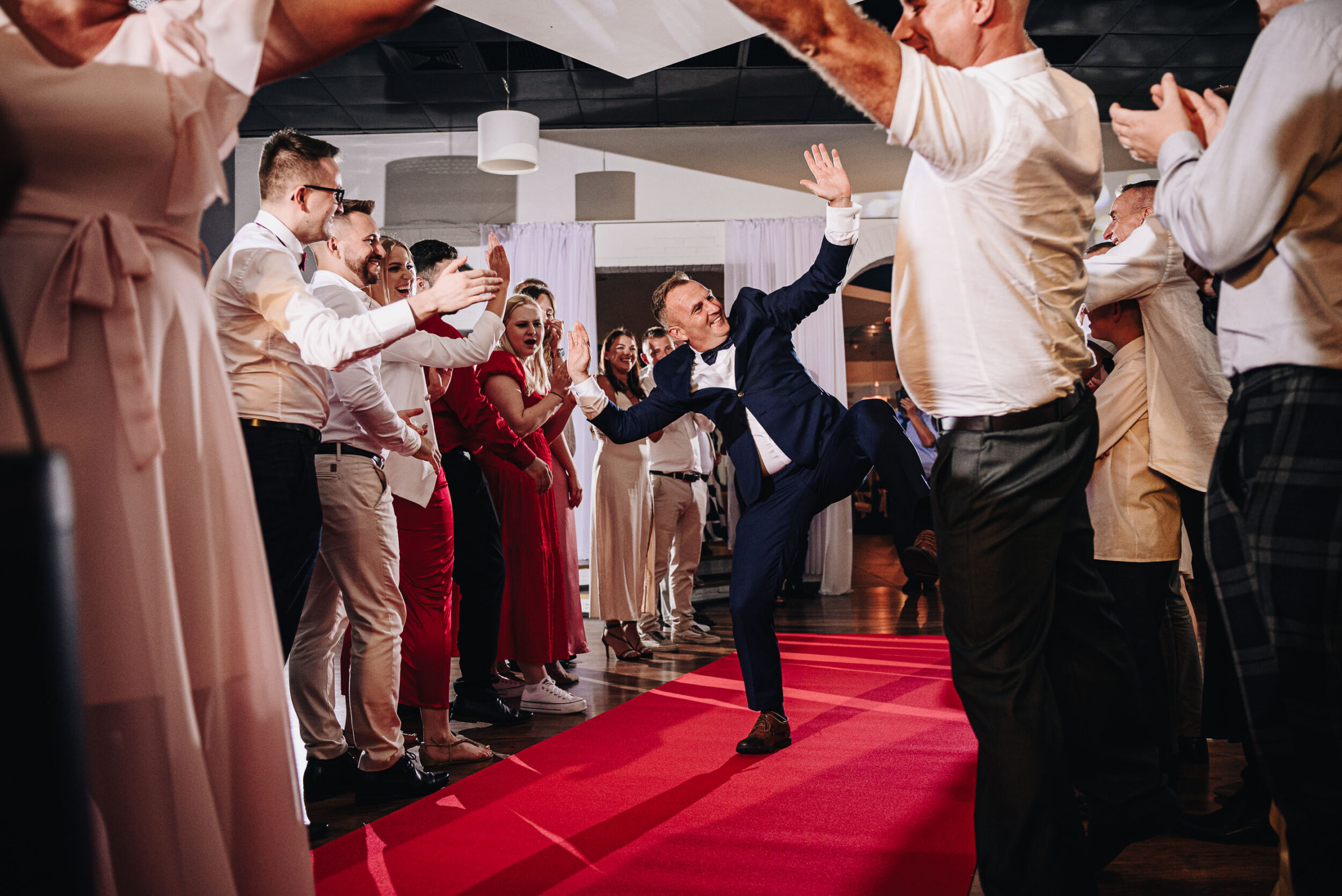 Uśmiechnięty mężczyzna w garniturze wykonuje dziwną pozycję na czerwonym dywanie. Po bokach stoją goście weselni, którzy przybijają z nim piątki.