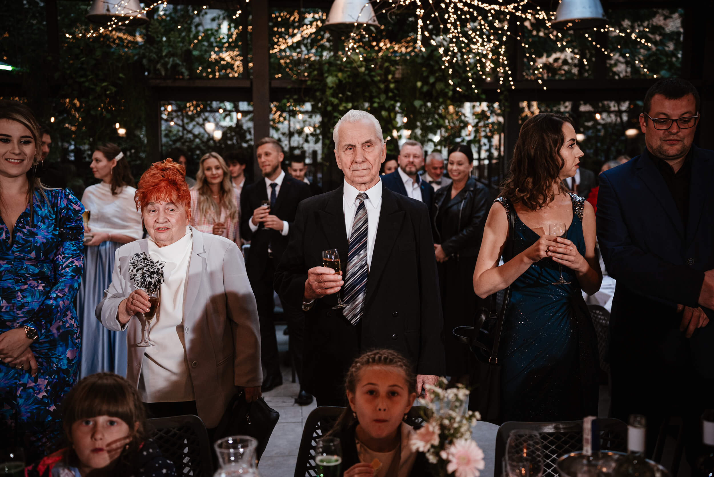 Elegancki starszy mężczyzna trzyma kieliszek wina. Obok stoją inni goście weselni.