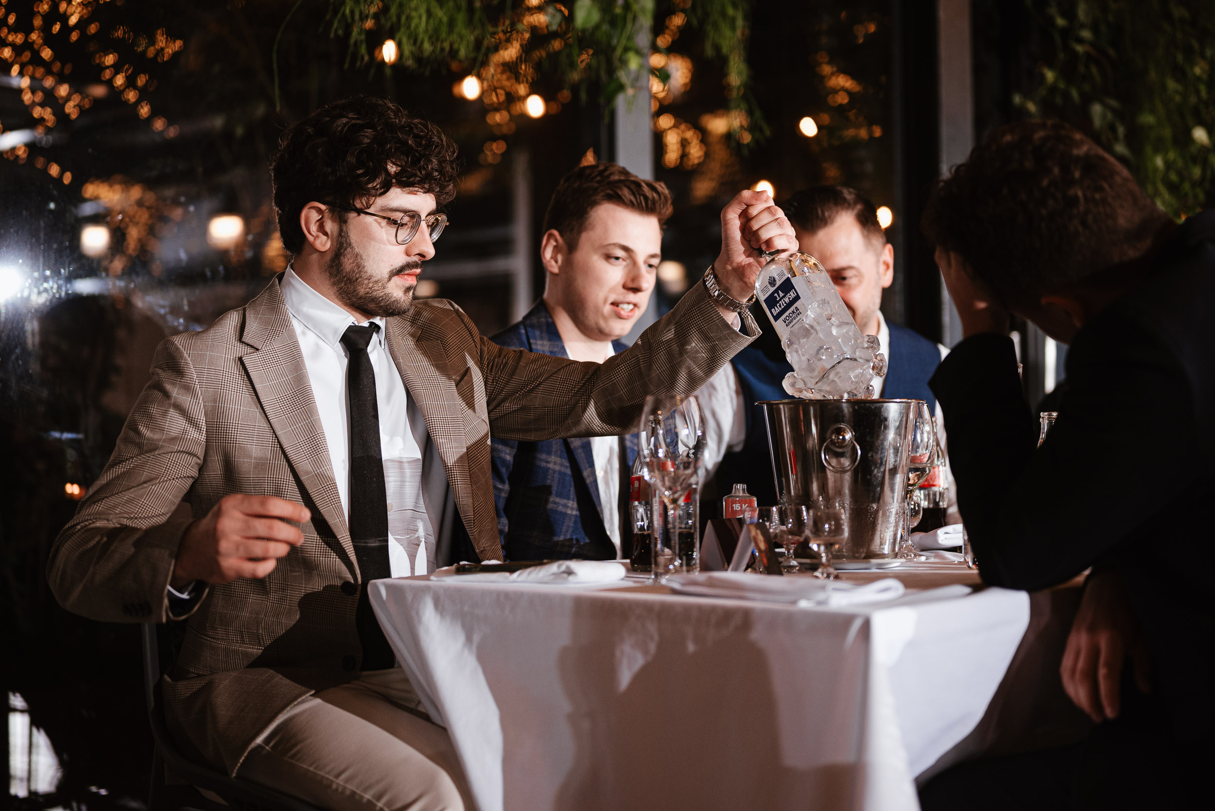 Gość weselny w beżowej marynarce i okularach wyciąga zamarzniętą butelkę wódki z wiaderka. Obok siedzą inni mężczyźni.